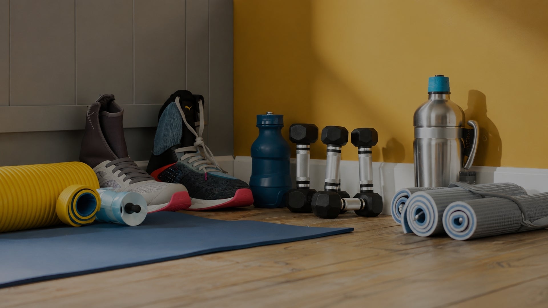 A gym workout setup with a blue exercise mat, gray and white sneakers, dumbbells, a blue water bottle, a stainless steel water bottle, and rolled-up resistance bands on a wooden floor against a yellow and gray wall.
