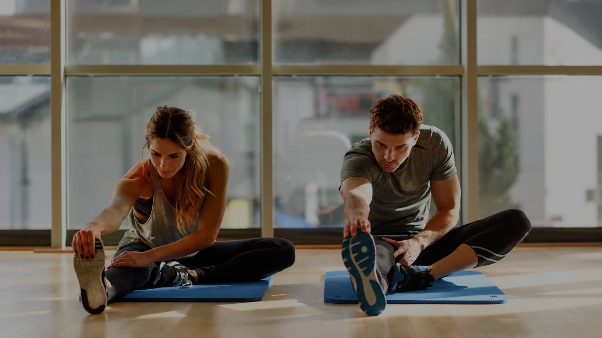 A man and a woman stretch on blue exercise mats in a bright gym with large windows.