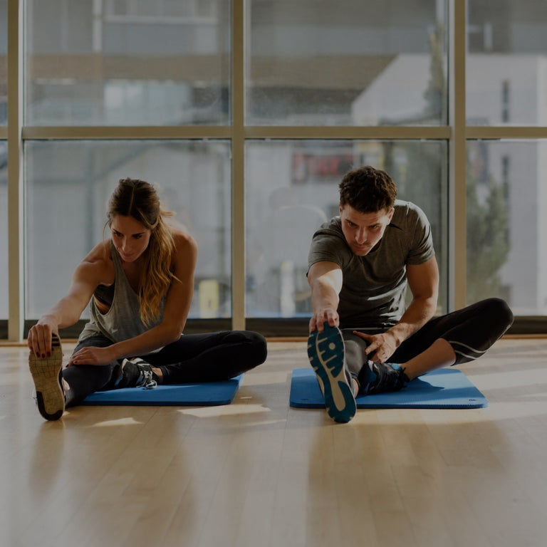A man and a woman stretch on blue exercise mats in a bright gym with large windows.